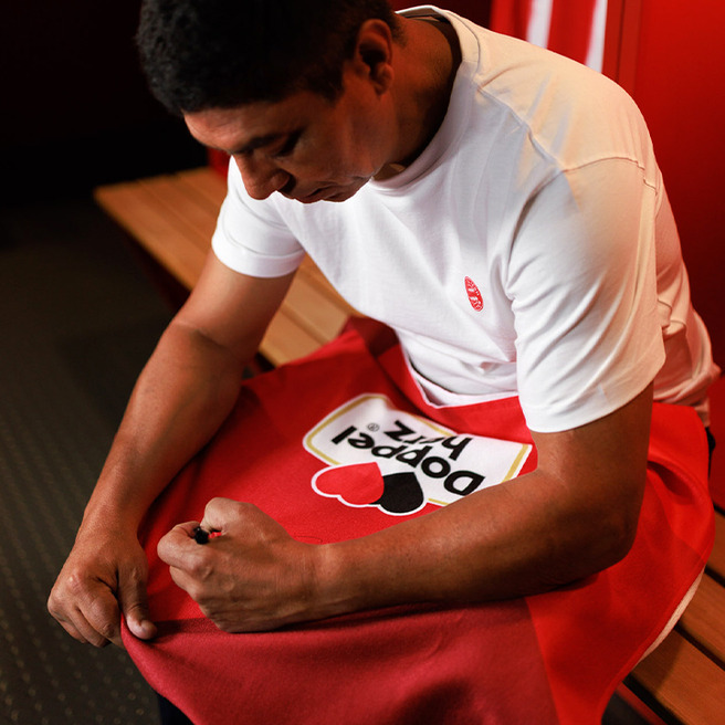 A man in a white shirt sits in a locker room, focused on adjusting a red and white item, likely a towel or garment with a heart design. He appears concentrated, with wooden benches and locker elements in the background.