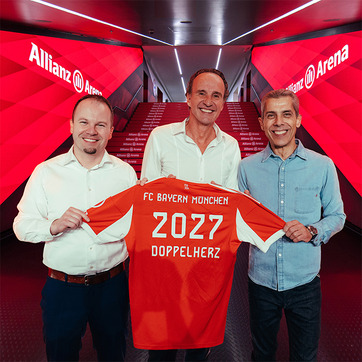 Three men stand together in a hallway of the Allianz Arena, holding up an orange FC Bayern München jersey. The jersey features the year "2027" and the word "DOPPELHERZ" on the back. The background highlights the team’s branding and colors.