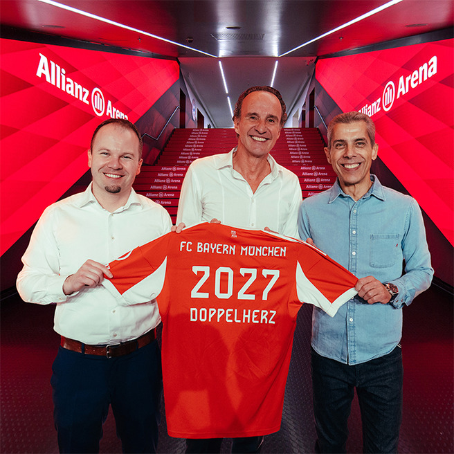 Three men stand together in a hallway of the Allianz Arena, holding up an orange FC Bayern München jersey. The jersey features the year "2027" and the word "DOPPELHERZ" on the back. The background highlights the team’s branding and colors.