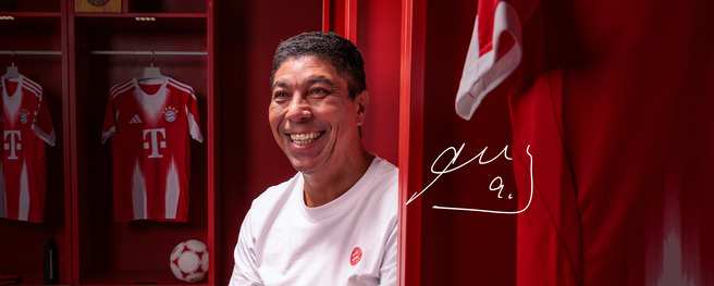 A smiling man sits in a red locker room, surrounded by FC Bayern Munich jerseys and a soccer ball. The walls are painted red, and a shirt hangs nearby. The image conveys a sense of joy and connection to football. A signature is visible in the corner.