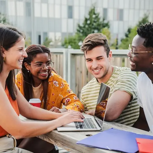 Four young adults sit together at an outdoor table, collaborating on a laptop. They appear engaged and happy, with one individual holding a coffee cup. Bright clothing and a casual setting suggest a friendly and relaxed atmosphere.