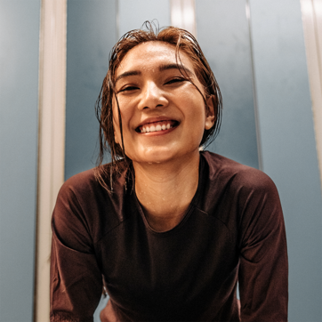 A smiling woman with wet hair leans forward, exuding joy and enthusiasm. She wears a dark athletic top, and the background features light blue walls, suggesting an energetic and active environment, possibly after a workout or swim.