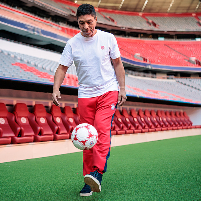 A person in a white t-shirt and red pants is skillfully juggling a soccer ball while standing on a green field. The background features empty stadium seats, emphasizing the sports environment.