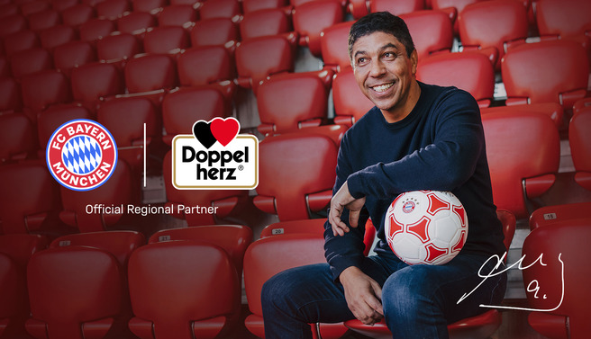 شخص مبتسم يجلس على مقاعد ملعب كرة قدم ويحمل كرة قدم، مع شعار نادي بايرن ميونيخ وشعار شريك رسمي.