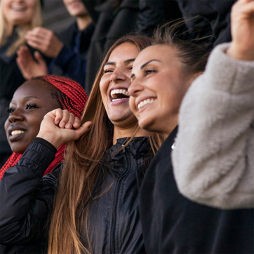 A group of three women express joy and excitement, smiling widely and laughing together. They are dressed in casual jackets, with one woman raising her hand in a celebratory gesture, while others appear engaged in a lively moment, suggesting a fun social atmosphere.