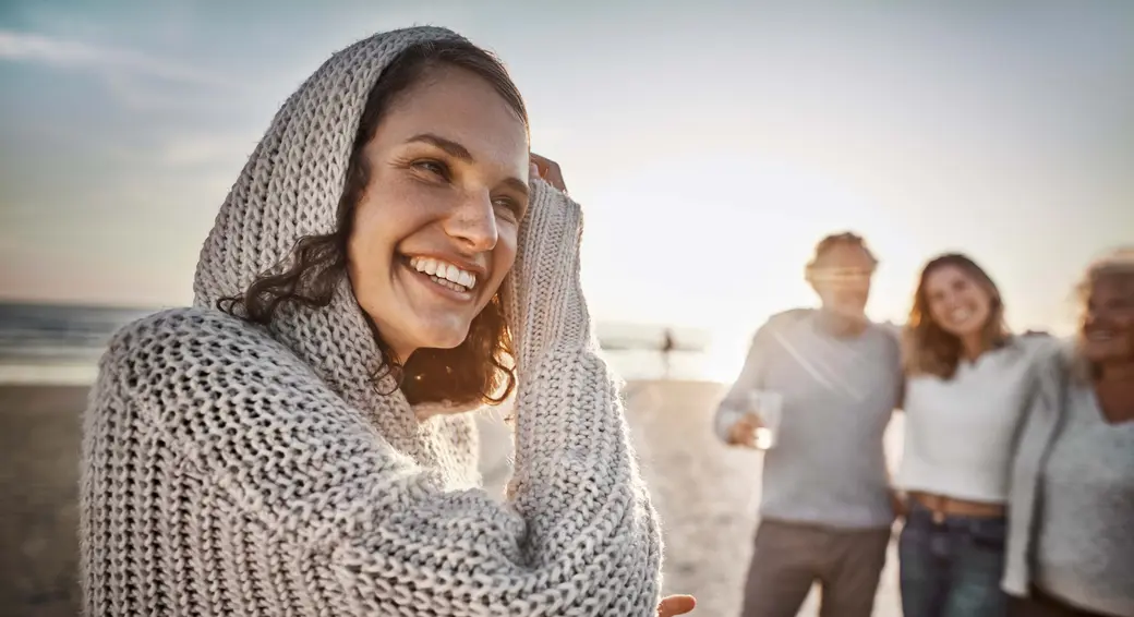 A smiling woman in a cozy, light-colored sweater stands on a beach, playfully pulling her hood over her head. The sun sets in the background, casting a warm glow, while three other people appear slightly blurred, enjoying the moment together.
