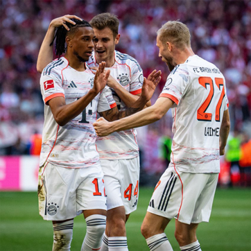 Three soccer players from Bayern Munich celebrate a goal on the field. One player, showing excitement, raises his hands, while another is smiling and extending a hand in celebration. The third player is lightly touching another player's head in a gesture of camaraderie.